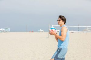 A young man in sunglasses holding a volleyball on a sunny beach, ready to play.