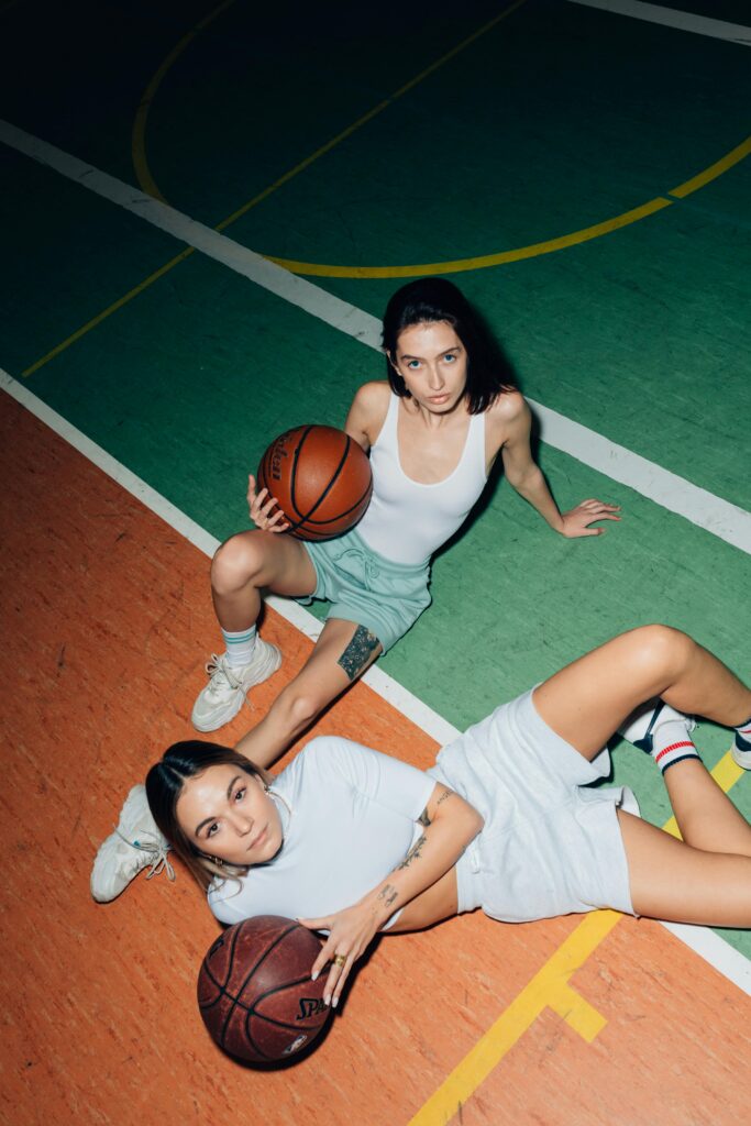Two women sitting on a basketball court holding basketballs, viewed from above.